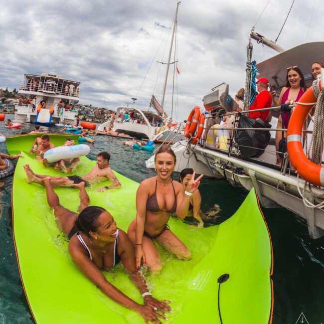A group of people in swimsuits enjoys a party on a large green floating mat and boats on the water. One woman in the center flashes a peace sign while others smile and play. The sky is cloudy, and boats are in the background.