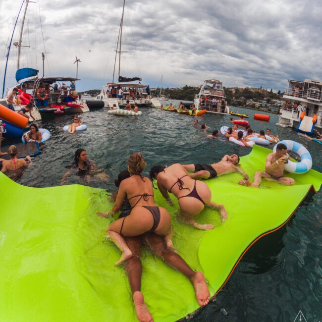 A group of people in swimwear relax and play on a large green floating mat in the water, surrounded by yachts and boats under a cloudy sky, enjoying a lively day party.