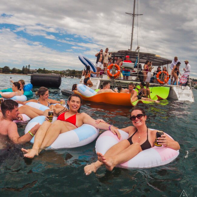 People relaxing on colorful inflatable tubes in the water near a boat, holding drinks and smiling; more people are on the boat, enjoying the scene under a cloudy sky.