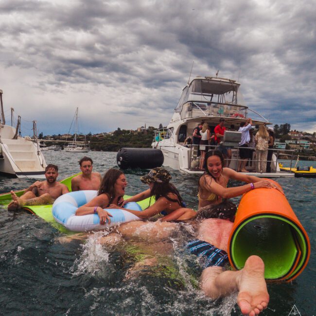 A group of young adults laugh and play on colorful floaties in the water near anchored boats under a dramatic cloudy sky. Some people watch from the boats while others enjoy the fun in the water.