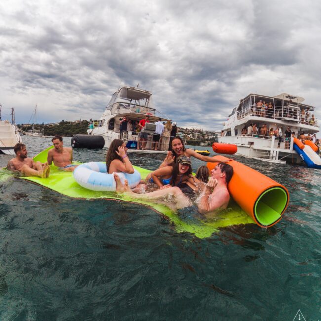 A group of young adults relax, laugh, and have fun on colorful floaties in the water near anchored party yachts, with more people visible on the boats under a cloudy sky.