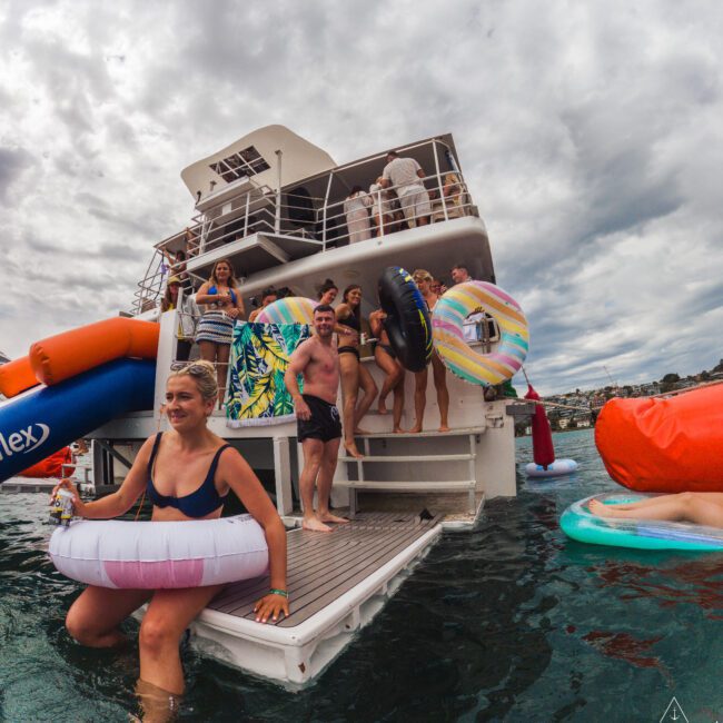 A group of people in swimsuits enjoy a boat party, some holding colorful inflatable rings and relaxing in the water, while others stand on the boat’s deck under a cloudy sky.