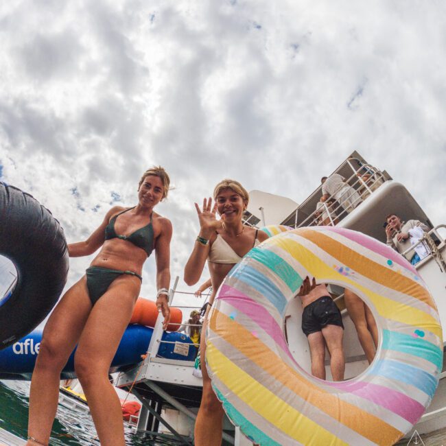 Two women in swimsuits stand on a docked boat, smiling and holding colorful pool floats. One waves at the camera. The sky is cloudy, and another person is stepping up a ladder in the background.