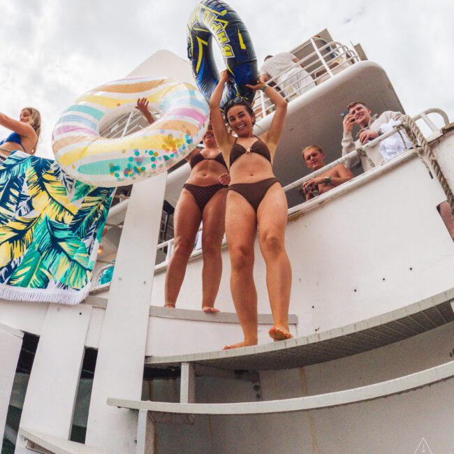 Two women in bikinis stand on a boat’s edge holding colorful pool floats above their heads, smiling and ready to jump, while people watch from the boat’s deck under a cloudy sky.