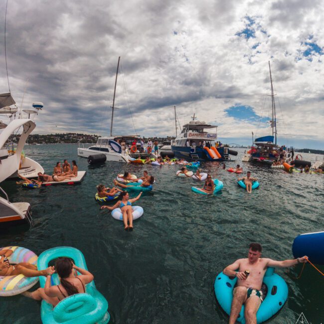People relax on colorful floaties in the water near several anchored boats under a cloudy sky. Others are sitting on the boats, and the scene looks lively and social, suggesting a boat party or gathering.