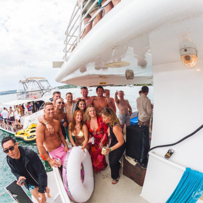 A group of people in swimwear are smiling and posing together on a boat, holding drinks, with water, jet skis, and another boat visible in the background on a sunny day.