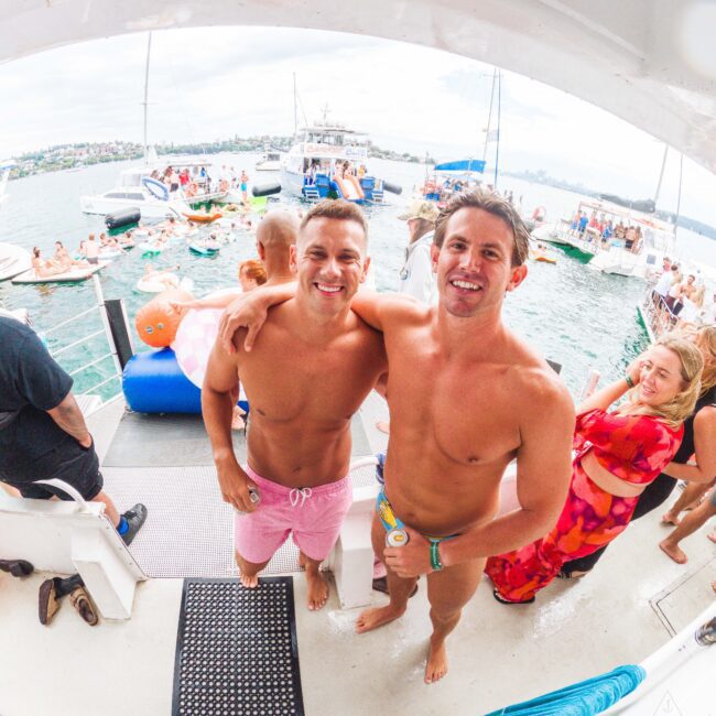 Two smiling men in swim trunks stand arm-in-arm on a boat, surrounded by people in swimwear. In the background, boats and paddleboards float on the water under a cloudy sky, and a woman in a red dress looks on.