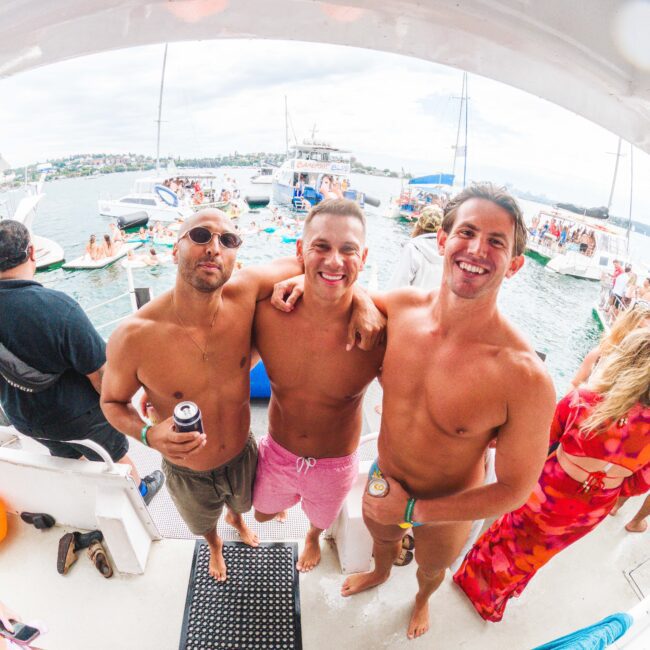Three smiling men in swim trunks stand arm-in-arm on a boat, enjoying a sunny day. Boats and people are visible in the water and background, suggesting a festive, social atmosphere.