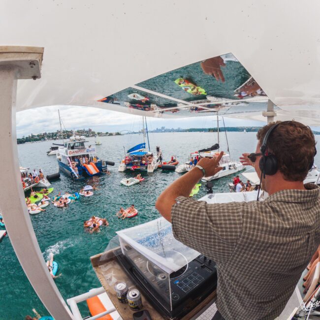 A DJ wearing headphones plays music on a boat, overlooking a lively gathering of people on floats and boats in the water, with city buildings visible in the distance.
