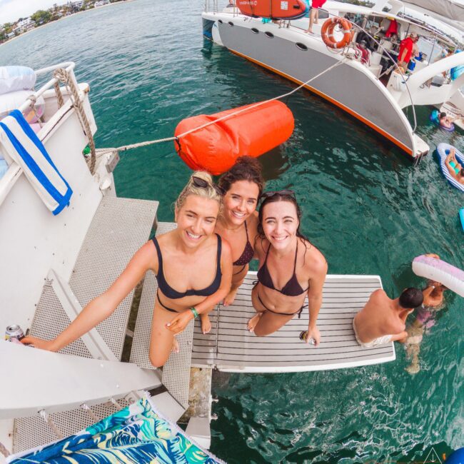 Three smiling women in swimsuits stand on a boat’s swim platform, posing for a photo with the ocean and another boat in the background. People relax nearby and colorful towels and floats are visible.