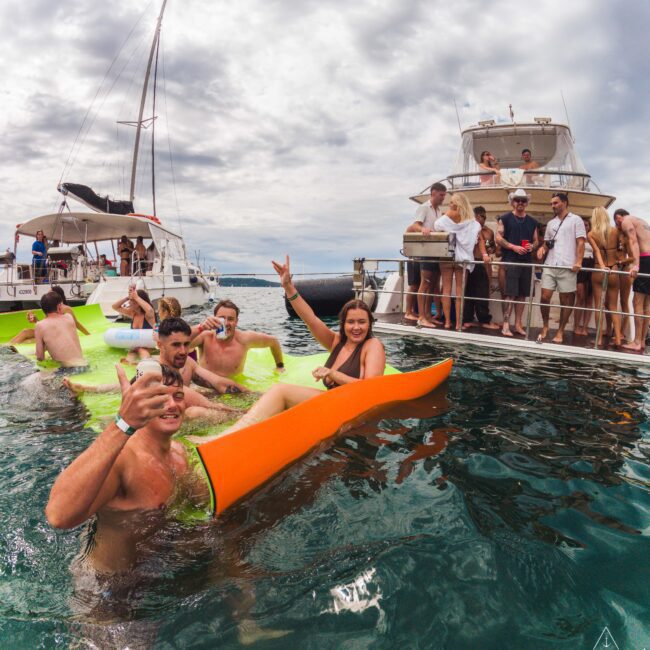 A group of people relax and have fun on floaties in the sea near yachts, with more people socializing on the boats. Everyone appears to be enjoying a lively party atmosphere under a cloudy sky.