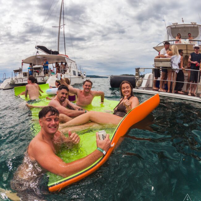 A group of young adults smile and relax on colorful floaties in the ocean between two boats during a cloudy day, with others socializing on the boats in the background.