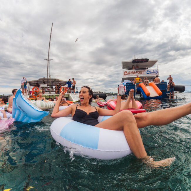 A woman in a black swimsuit laughs while floating on a blue and white inflatable ring in the ocean near a party boat, surrounded by other people and floats under a cloudy sky.