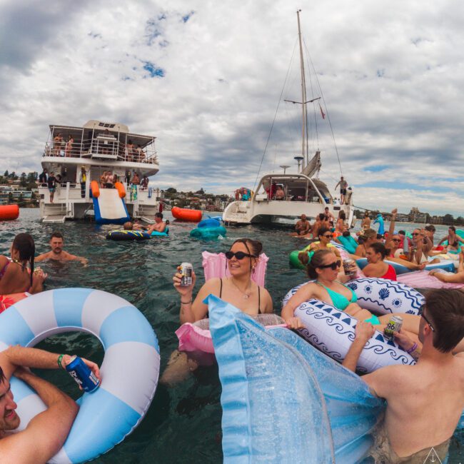A group of people relax on colorful inflatables and floaties in the water near two anchored boats under a cloudy sky, holding drinks and enjoying a lively social gathering.