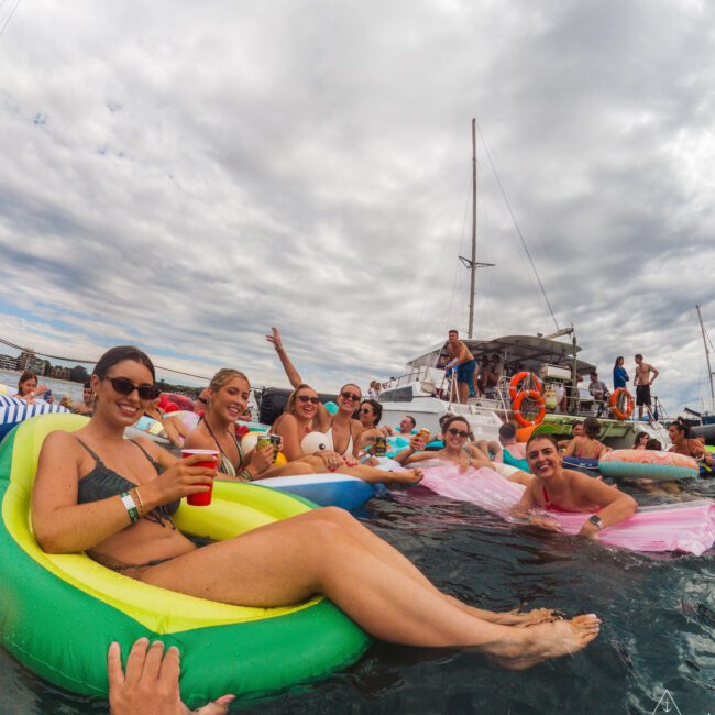 A group of young adults relax on colorful inflatables in the water, smiling and enjoying drinks near a docked yacht under a cloudy sky. Some people are on the yacht, and the atmosphere is lively and festive.