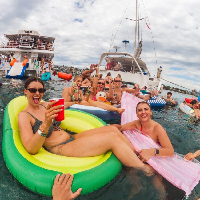 A group of people relax and smile on colorful pool floats in the water near yachts, holding drinks and posing for the camera at a lively boat party on a partly cloudy day.