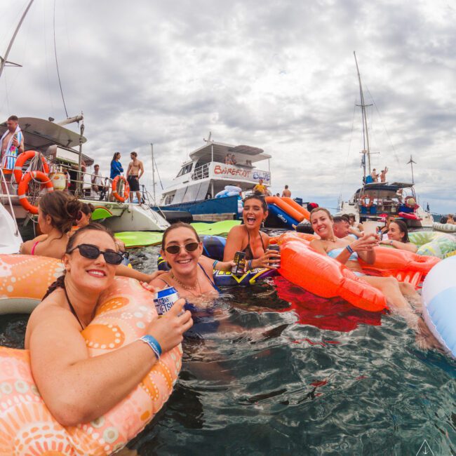 A group of smiling people relax on colorful pool floats in the water near several boats on a cloudy day, enjoying drinks and socializing at a lively outdoor gathering.