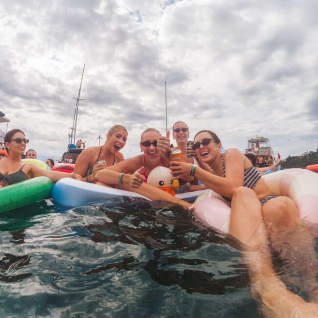 A group of smiling women in swimsuits float on inflatable tubes in the water, holding drinks and posing for a photo. Sailboats and yachts are visible in the background under a cloudy sky.