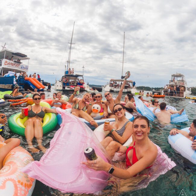 A group of people relaxing on colorful pool floats, holding drinks and smiling in the water, with boats anchored nearby under a cloudy sky.