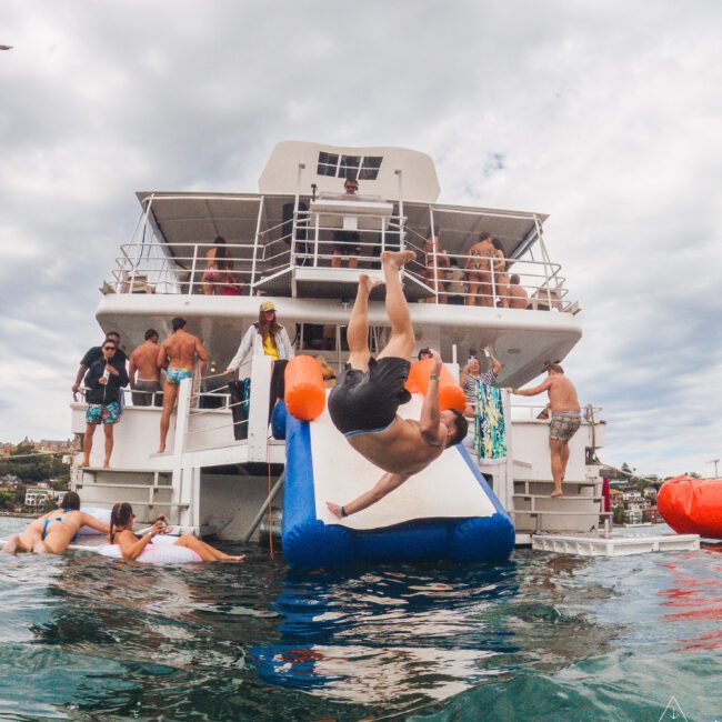 A man slides off an inflatable slide attached to a docked boat, mid-air above the water. People on the boat and in the water watch, enjoying a lively day under a cloudy sky.