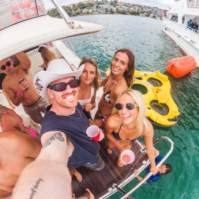 A group of smiling friends in swimwear pose for a selfie on a boat, holding drinks. Behind them are floating rafts, clear blue water, and other boats, with a shoreline visible in the distance.
