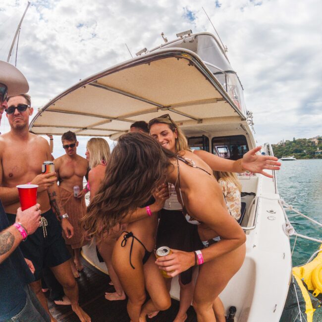 A group of young adults in swimsuits with pink wristbands enjoy drinks and laugh together on the deck of a boat under a partly cloudy sky, anchored near the shore.