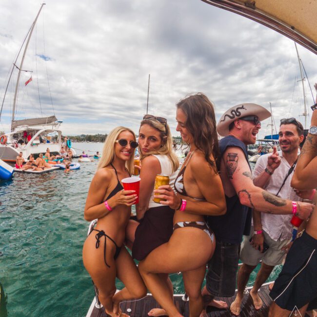 A group of people in swimsuits and party attire stand on a boat, holding drinks and posing for a photo. Other boats and people are visible in the water under a cloudy sky.