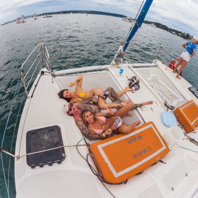 Three people in swimsuits relax and smile on the deck of a sailboat, lounging near orange safety containers labeled "Captain of Joy," with water and other boats visible in the background.