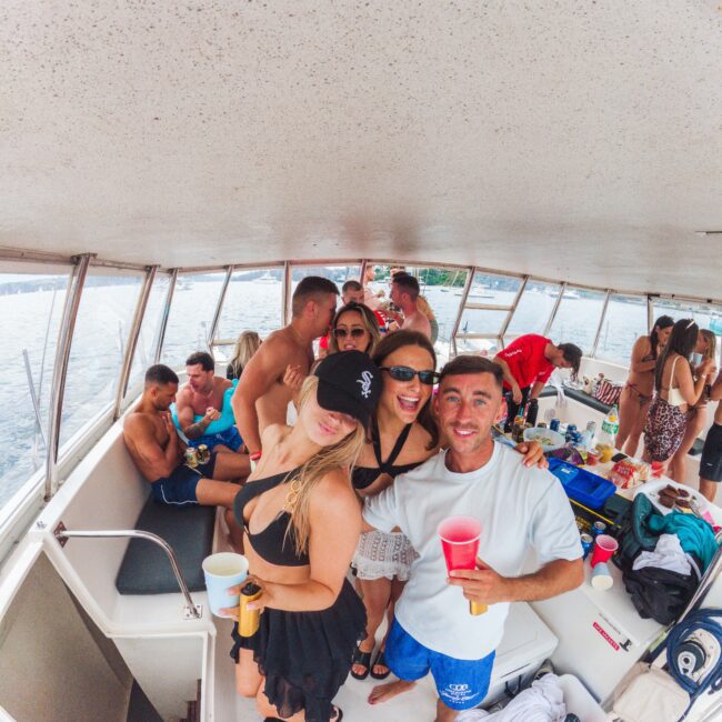 A group of young adults in swimsuits smiles and poses with drinks on a boat, surrounded by others socializing and enjoying a sunny day on the water. The mood is festive and relaxed.