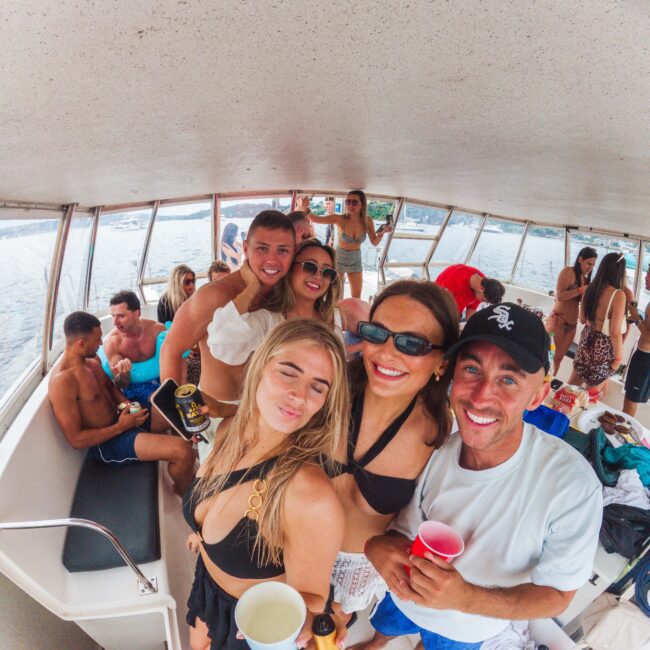 A group of young adults in swimsuits smile and pose with drinks on a boat. More people are in the background, enjoying the party near the water. The atmosphere is lively and festive.