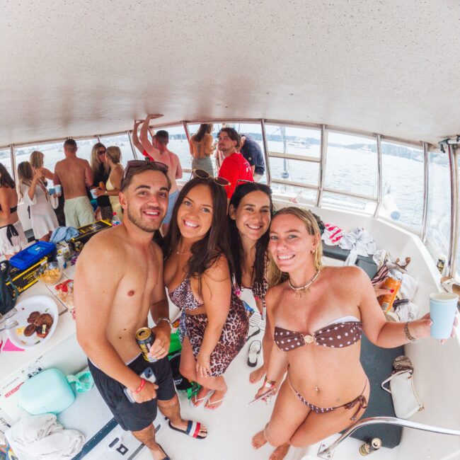 A group of young adults in swimsuits smile and pose for a photo on a boat, surrounded by drinks and snacks, with more people socializing in the background near large windows showing water outside.