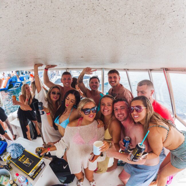 A group of smiling young adults in swimsuits and cover-ups pose closely together on a boat, holding drinks, with water and more people visible outside the windows in the background.