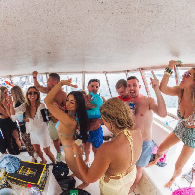A group of young adults in swimwear enjoy a lively party on a boat, dancing, laughing, and holding drinks. The mood is festive, with people posing and having fun near large windows overlooking the water.