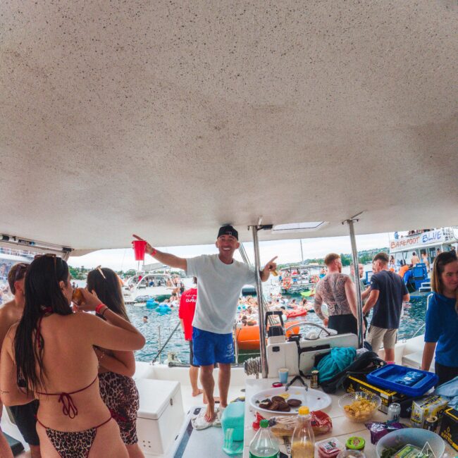 A group of people gather on a boat with food and drinks on a table. One person stands holding a red cup, smiling. Other boats and people are visible in the water in the background on a sunny day.