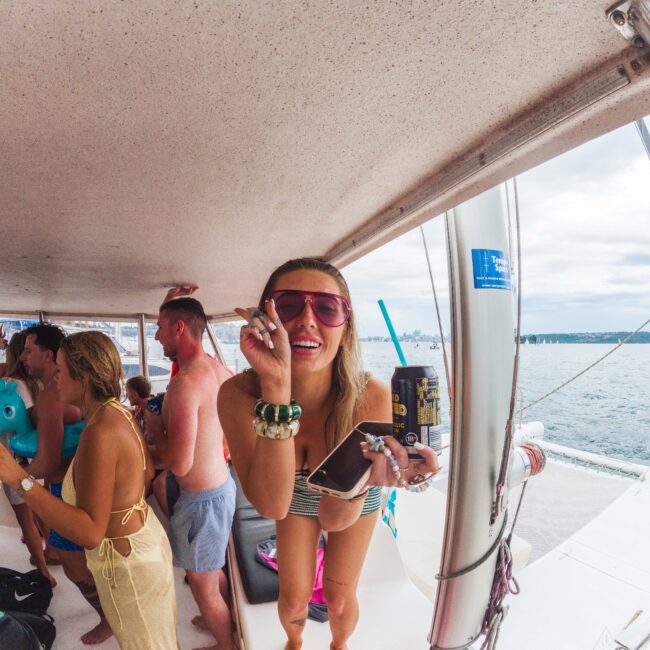 A woman in sunglasses smiles and gestures peace sign while holding a drink on a boat crowded with people in swimsuits; the sea and coastline are visible in the background.