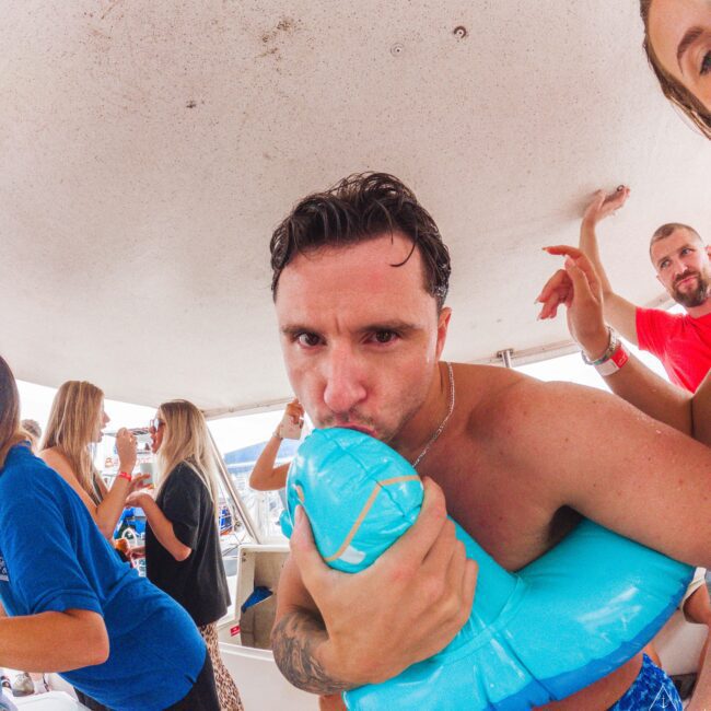 A man in swim trunks kisses a blue inflatable pool toy at a lively party on a boat, surrounded by people laughing, talking, and enjoying the event. The atmosphere is casual and festive.