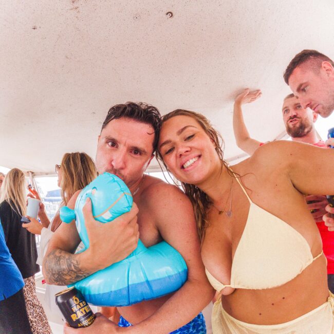 A man hugs an inflatable blue dinosaur and poses with a smiling woman in a cream bikini top. They are on a boat with several other people in swimsuits, enjoying a lively, casual atmosphere.