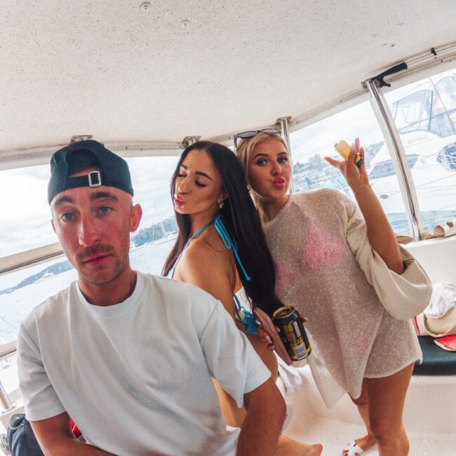 Three young adults pose on a boat; a man in a white t-shirt and hat stands beside two women in swimsuits, one with a drink and the other with a can. All are smiling and making playful expressions. Water and boats are visible outside.
