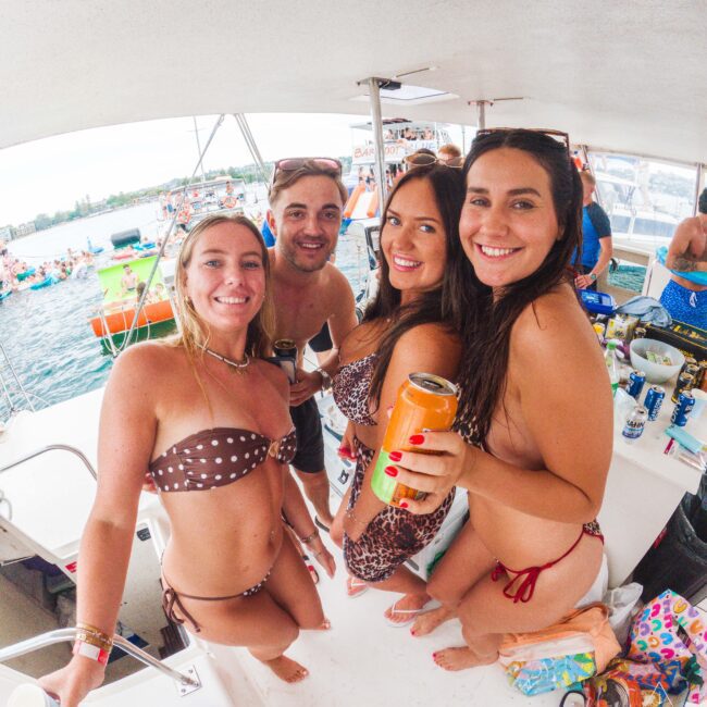 Four smiling people in swimsuits hold drinks on a boat, surrounded by water and other boats filled with people in the background, enjoying a lively, sunny day.