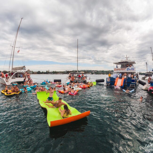 People relax on colorful floats and inflatables in the water near anchored boats under a cloudy sky. Some people sit on a large green floating mat, while others swim or lounge around the boats.