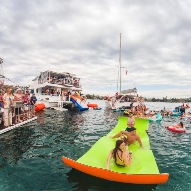 A group of people enjoy a party on boats and floating mats in the water. Some relax on a large green float, while others socialize on the boats and swim nearby under a cloudy sky.
