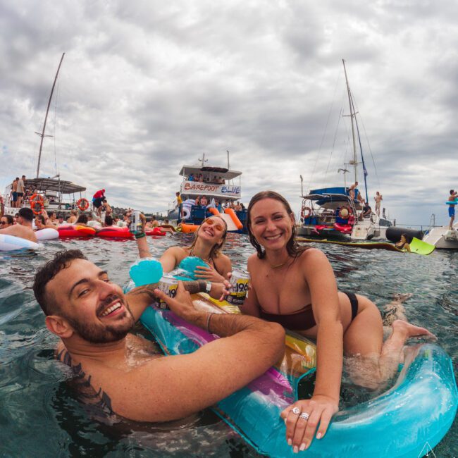 Three people in swimsuits smile and relax on colorful inflatables in the water, with boats and other people partying in the background under a cloudy sky.