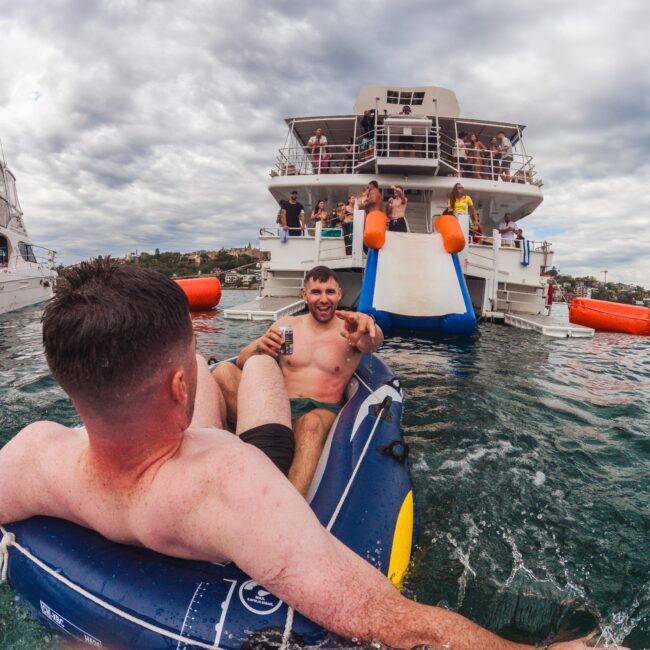 Two men in swim trunks relax on inflatables in the water near a large party boat, which has a slide and several people on board. The sky is cloudy and people are enjoying the scene.