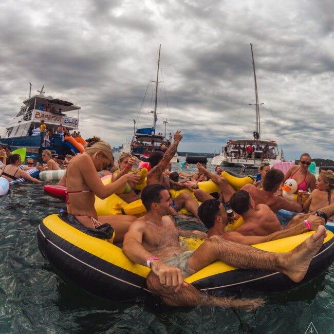 A group of people in swimsuits relax and have fun on a large yellow inflatable raft in the water, with boats and other floaters nearby under a cloudy sky.