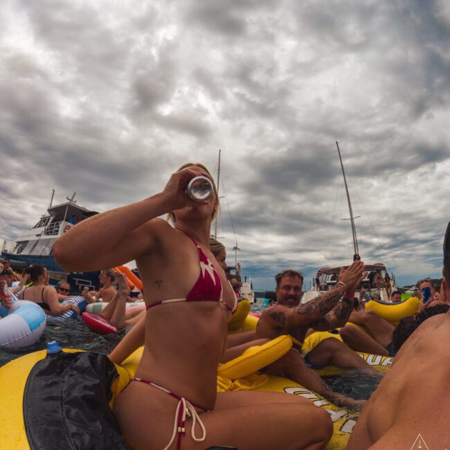 A group of people in swimsuits relax on yellow inflatable tubes, drinking and socializing in the water near boats under a cloudy sky. The scene is lively and festive.