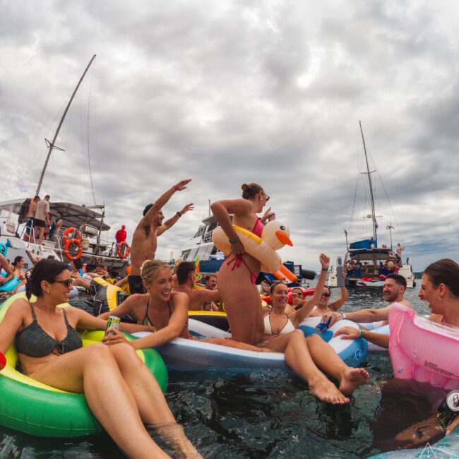 A group of people in swimsuits relax and have fun on colorful pool floats in the ocean near anchored boats, under a cloudy sky. One person wears an inflatable duck around their waist.