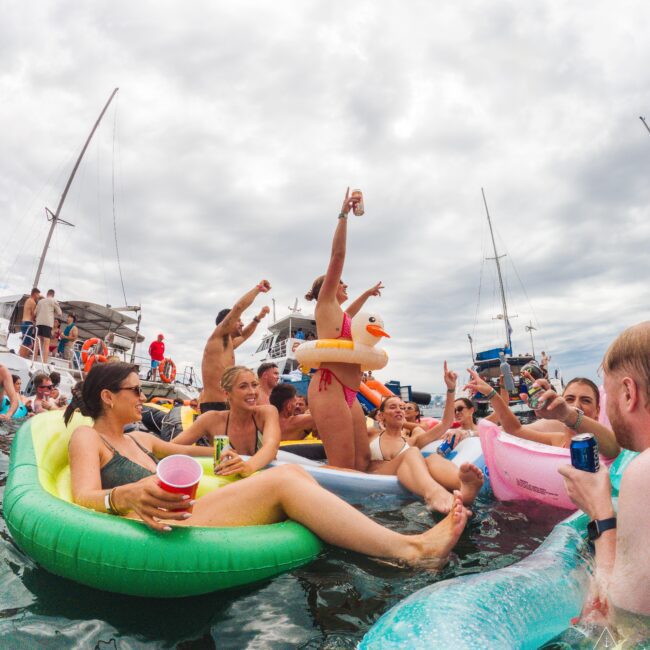 A group of people enjoy a lively party on inflatable floats in the water, raising drinks, laughing, and celebrating near docked sailboats under a cloudy sky.