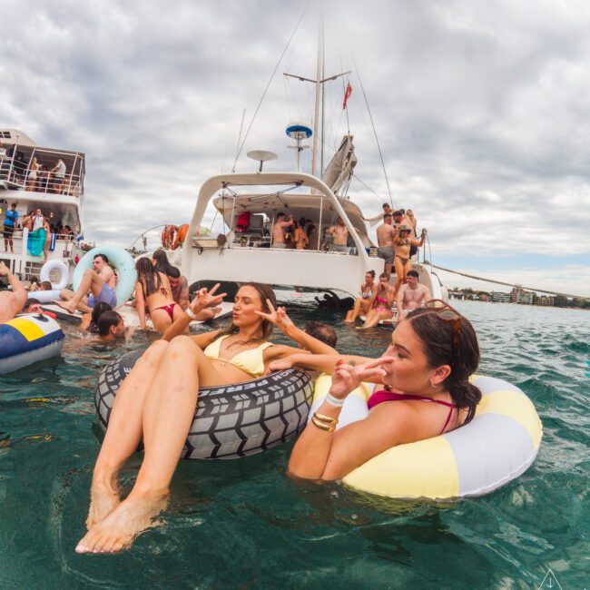 Two women relax on inflatable rings in the water near a boat packed with people, all enjoying a lively party under a cloudy sky. Several others float nearby, creating a festive atmosphere.