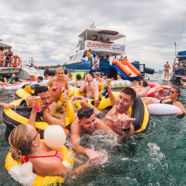 A group of young adults relax on colorful floaties in the water, holding drinks and smiling, near a large yacht labeled “Barefoot Blue” on a cloudy day. Other boats and people are seen in the background.