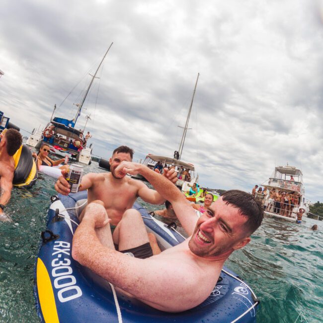 Two smiling men in swim trunks sit in a small inflatable raft on the water, surrounded by boats and people under a cloudy sky. One man gives a thumbs-up while others enjoy the lively, social atmosphere.